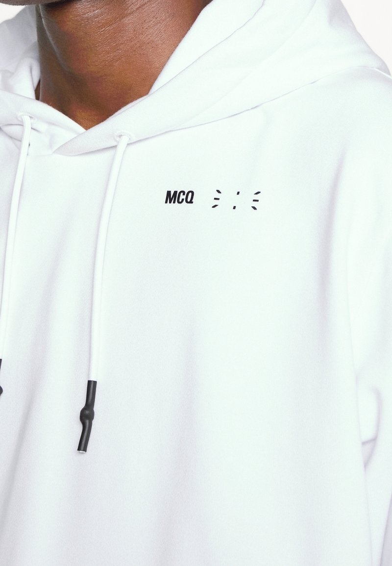 White hoodie with black graphic text "MCQ" and line accents. Features drawstring hood and textured fabric. Pullover design, relaxed fit.