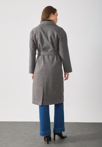 Grå ullfrakk med belte i midjen, lange ermer og skjært krage. Bakdelen har en sentral slisse og når til midt på leggen.