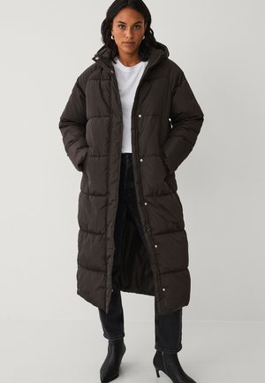 Lange, gevoerde donkerbruine jas met een capuchon, voorzien van een rits- en drukknoopsluiting, zijzakken en een gestikt ontwerp. Gecombineerd met zwarte jeans en laarzen.