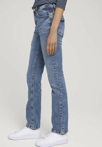 Jean en denim bleu clair avec une taille ajustée, jambes légèrement évasées et décoloration subtile. Associés à des baskets blanches pour un contraste.