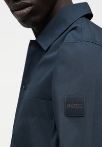 Κοντινό πλάνο ενός σκουρόχρωμου ατόμου που φοράει σκούρο μπλε πουκάμισο με τετράγωνο έμβλημα BOSS στον άνω μανίκι.