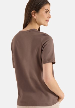 Femme portant une chemise marron à manches courtes avec un bord en dentelle œillet sur les manches, debout, la main dans la poche, tournée de profil.