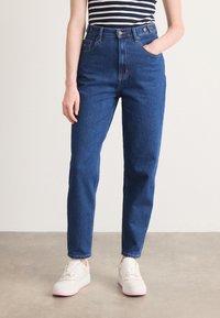 Høyt midje blå jeans laget av denim med rett passform, fem-lomme-design og knappelukking foran, kombinert med hvite joggesko.