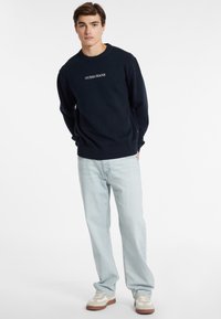 Tengerészkék crewneck pulóver bordás mandzsettával és aljjal, rajta a "GUESS JEANS" logó. Világoskék farmernadrággal és fehér sportcipővel párosítva.