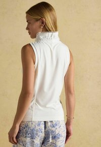 Bílý bezrukávový top s vysokým volánovým límcem, měkká tkanina, slim fit a boční švy. Nositý s vzorovanými modro-bílými kalhotami.