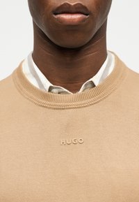 Gros plan d'une personne portant un pull beige par-dessus une chemise rayée à col, avec le logo "HUGO" brodé sur le pull.
