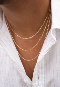 Goldene Kettenhalsketten in verschiedenen Längen übereinander gelegt über einem nackten Oberkörper, mit einem weißen Hemd mit Kragen, das subtile Streifen und Knopfdetails aufweist.