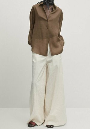 Person wearing a loose brown button-up shirt, white wide-leg pants, black sandals, and a black neck scarf, standing against a plain background.