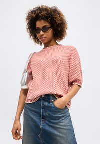 Oseba z kodrastimi lasmi, ki nosi črna sončna očala, roza teksturiran pulover s kratkimi rokavi, jeans krilo in srebrno torbico čez ramo.