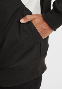 Svart jakke med hvite detaljer, med en sidelomme og ribbestrikkede mansjetter. Stoffet ser glatt ut med en matt overflate.