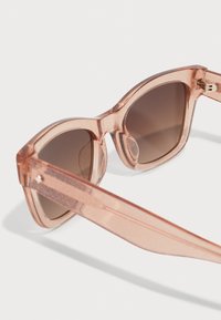Sunglasses with a transparent peach frame, brown tinted lenses, and a textured finish. Straight arms with subtle gold hardware accents.