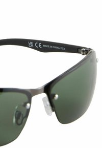 Ochelari de soare cu ramă din plastic negru, lentile cu nuanță verde, pernuțe de nas ajustabile și accente metalice. Marcajele de pe tâmple includ CE, UKCA și "FABRICAT ÎN CHINA."