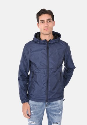 Giacca impermeabile blu navy con cappuccio, chiusura con zip, polsini elasticizzati e tasche laterali. Presenta una texture liscia e lucida. Indossata con jeans in denim.