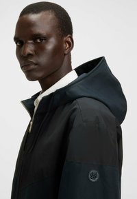 Chaqueta negra con capucha y textura suave, con un panel contrastante y una insignia redonda en la manga. Cierre de cremallera en la parte frontal.