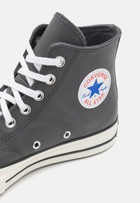 Svetlošedé kožené vyšívané tenisky Converse s bielymi šnúrkami, okrúhlymi očkami, modrou hviezdou na patke a textúrovanou gumovou podrážkou s čiernymi akcentmi.