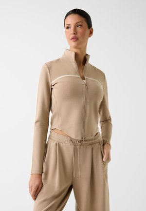 Donna che indossa una giacca beige con cerniera e pantaloni coordinati con coulisse, in piedi con una mano in tasca, che guarda di lato su sfondo bianco.