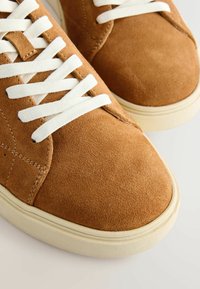 Gros plan sur des baskets en daim marron avec des lacets blancs et des semelles en caoutchouc blanc cassé sur un fond clair.