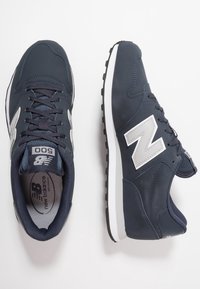 Zapatillas deportivas de color azul marino, hechas de material sintético, con un logotipo "N" plateado, cuello acolchado y suela blanca con agarre texturizado.
