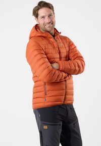 Oranje pufferjas met een capuchon heeft een gequilte ontwerp, een ritssluiting aan de voorkant en twee zijzakken. G gedragen met donkere broek.