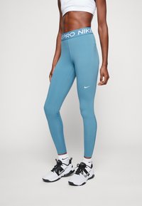 Leggings haute taille bleues avec une large bande élastique marquée "PRO NIKE" et un petit logo Nike blanc sur la jambe gauche. Conception en tissu extensible.