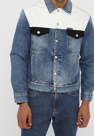 Kurtka dżinsowa z białym karczkiem i czarnymi kieszeniami z klapami, noszona z niebieskimi dżinsami przez stojącą osobę.