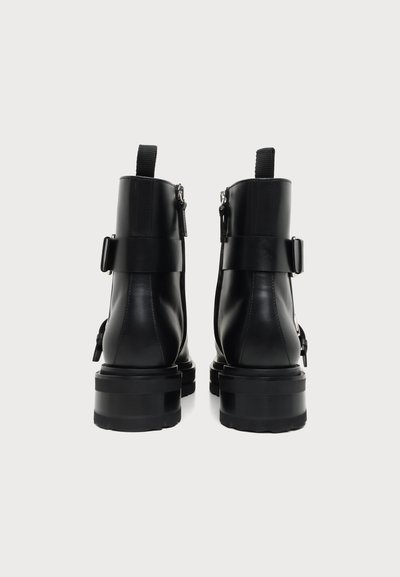Paire de bottines en cuir noir avec semelles épaisses, languettes à l'arrière, fermetures éclair latérales et sangles décoratives avec boucles, vue de dos.