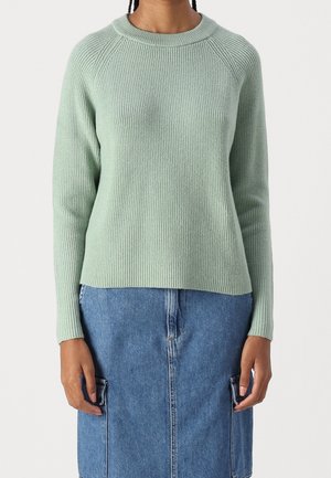 Persoon draagt een lichtgroene, geribbelde trui met lange mouwen en een blauwe denim rok met voorzakken, staand tegen een effen achtergrond.