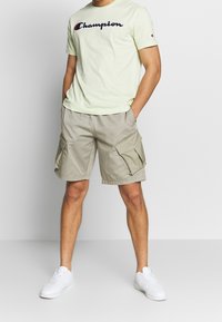 Svetle zelené bavlněné tričko s logem "Champion" v tmavě modré a červené barvě, spárované s béžovými cargo šortkami s klopovými kapsami. Bílé tenisky.