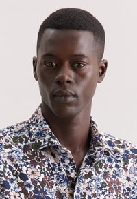 Camicia floreale con fondo bianco che presenta fiori blu, verdi e marroni. Stile con bottoni, collo a punta e tessuto testurizzato.