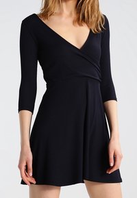Czarna, prążkowana sukienka o luźnym kroju z głębokim dekoltem w kształcie litery V i rękawami o długości trzy czwarte. Rozkloszowany dół sukienki sięga powyżej kolan. Gładka faktura, prosty design.