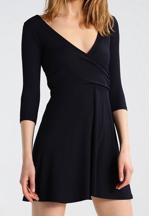 Mustan, ribattu kiertopuku, jossa on syvä V-pääkallo ja kolmen neljänneksen hihat. Valloittava helma ulottuu polven ylle. Sujuva pinta, yksinkertainen muotoilu.