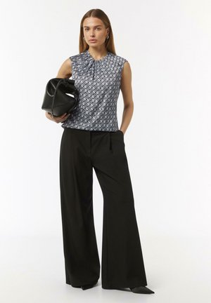 Ärmelloses, navy-blaues Top mit Muster, Kragen und Falten, kombiniert mit schwarzen, weit geschnittenen Hosen. Das Model hält eine schwarze Handtasche.