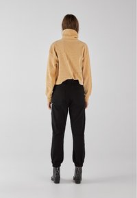 Pull turtleneck beige à côtes associé à un pantalon cargo noir avec des poignets fuselés. Le mannequin se tient de dos, portant des bottes noires.