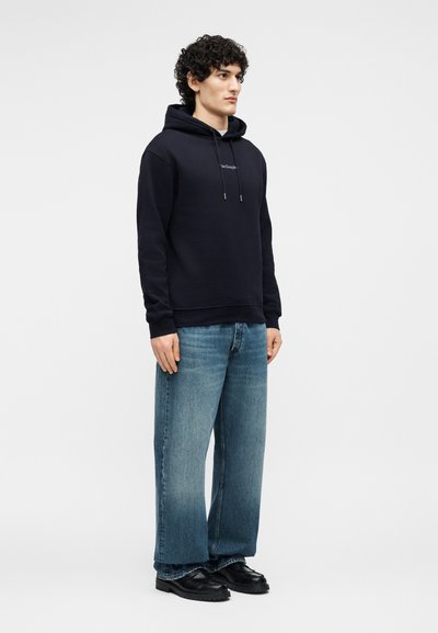 Homme aux cheveux bouclés portant un sweat à capuche noir, un jean bleu à jambes larges et des chaussures noires, debout devant un fond clair uni.