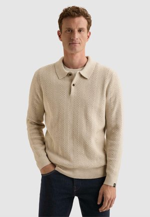 Hombre con cabello rizado que lleva un suéter de punto beige texturizado con cuello y tres botones, combinado con vaqueros azul oscuro.