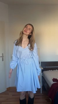 Νέα γυναίκα με μακριά ξανθά μαλλιά που φοράει ένα ανοιχτό μπλε φόρεμα και μαύρες μπότες στέκεται σε ένα υπνοδωμάτιο με ξύλινο πάτωμα και μπεζ τοίχους.