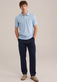 Hellblaues Poloshirt mit Kragen und einem Logo auf der Brust, kombiniert mit dunkelblauen Hosen. Die Schuhe sind beige mit einer strukturierten Oberfläche.