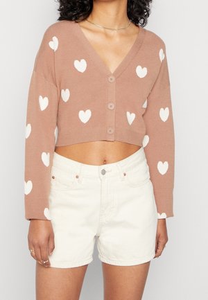 Vrouw draagt een cropped, bruine cardigan met knopen en witte hartpatronen, gecombineerd met hooggetailleerde witte denim shorts.