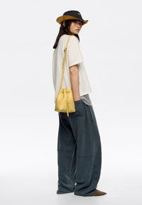 Helles gelbes Leder-Bucket-Bag mit Zugverschluss, quer über den Körper getragen; Modell trägt ein cremefarbenes T-Shirt und übergroße blau-graue Hose.