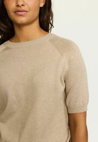 Maglione corto a maniche corte di colore beige, con collo rotondo e maniche a raglan, che mette in mostra una texture liscia e sottili dettagli di cucitura.