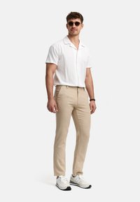 Camisa blanca de botones de manga corta, pantalones beiges, usando gafas de sol y zapatillas blancas. La camisa tiene un diseño relajado y una tela texturada.