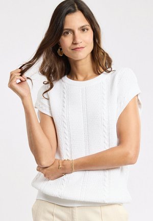 Femme aux longs cheveux bruns portant des boucles d'oreilles créoles dorées et un pull blanc à manches courtes en maille torsadée, tenant une mèche de cheveux.