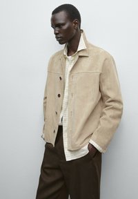 Beige Wildlederjacke mit Knopfleiste, zwei Taschen und einem darunter getragenen Hemd mit einem Karomuster in Hellgrün und Weiß.