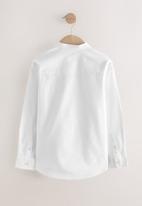 Chemise blanche à manches longues avec boutons, suspendue sur un cintre en bois contre un mur clair uni, vue de dos.