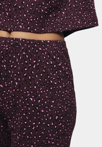 Fleksibel to-dels sæt i mørk burgunder, der inkluderer en cropped top og bukser. Stoffet har et sort og pink leopardprint mønster.