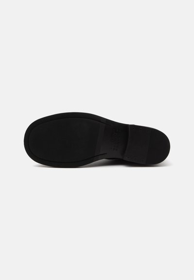 Semelle de chaussure habillée noire vue de dessous, montrant une surface lisse avec de fines rainures et un petit talon sur fond blanc uni.