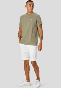 Olivengrønn bomulls T-skjorte med et trykt smilefjes-design på venstre bryst, kombinert med hvite bomullsshorts og hvite sneakers.