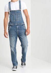 Overoles de mezclilla azul con tirantes ajustables, bolsillos frontales y un corte relajado. Se llevan con una camiseta blanca y zapatillas negras.