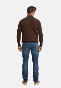 Hombre de pie de espaldas, vistiendo un suéter marrón, jeans azules y zapatos beige, contra un fondo blanco liso.