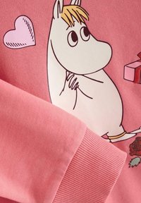 Roze sweatshirt met een Moomin-personage met geel haar, dat handen samenhoudt, tegen een lichtroze achtergrond met een hart en cadeaus.